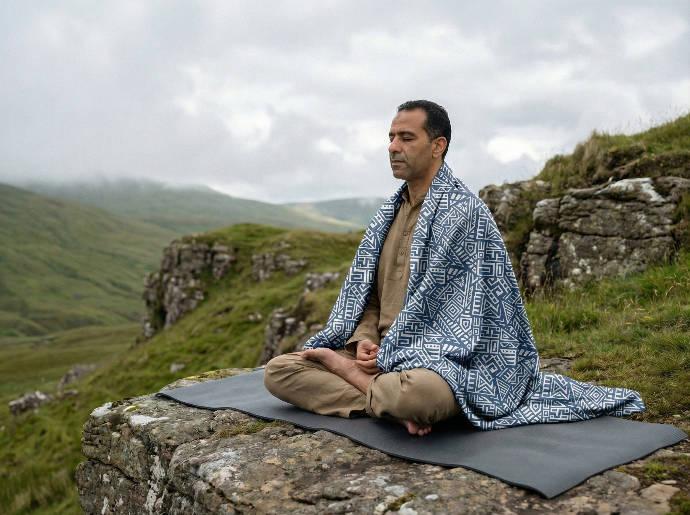 Man meditating outdoors on a yoga mat wearing a blue geometric patterned meditation shawl, practicing mindfulness in a misty mountain landscape.
