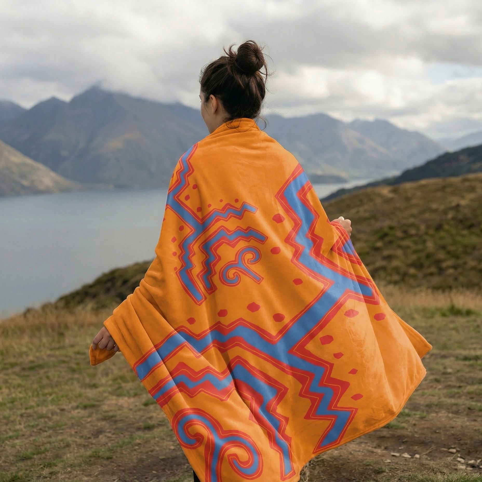 Person standing outdoors wearing an orange throw blanket featuring abstract geometric and tribal-inspired patterns.