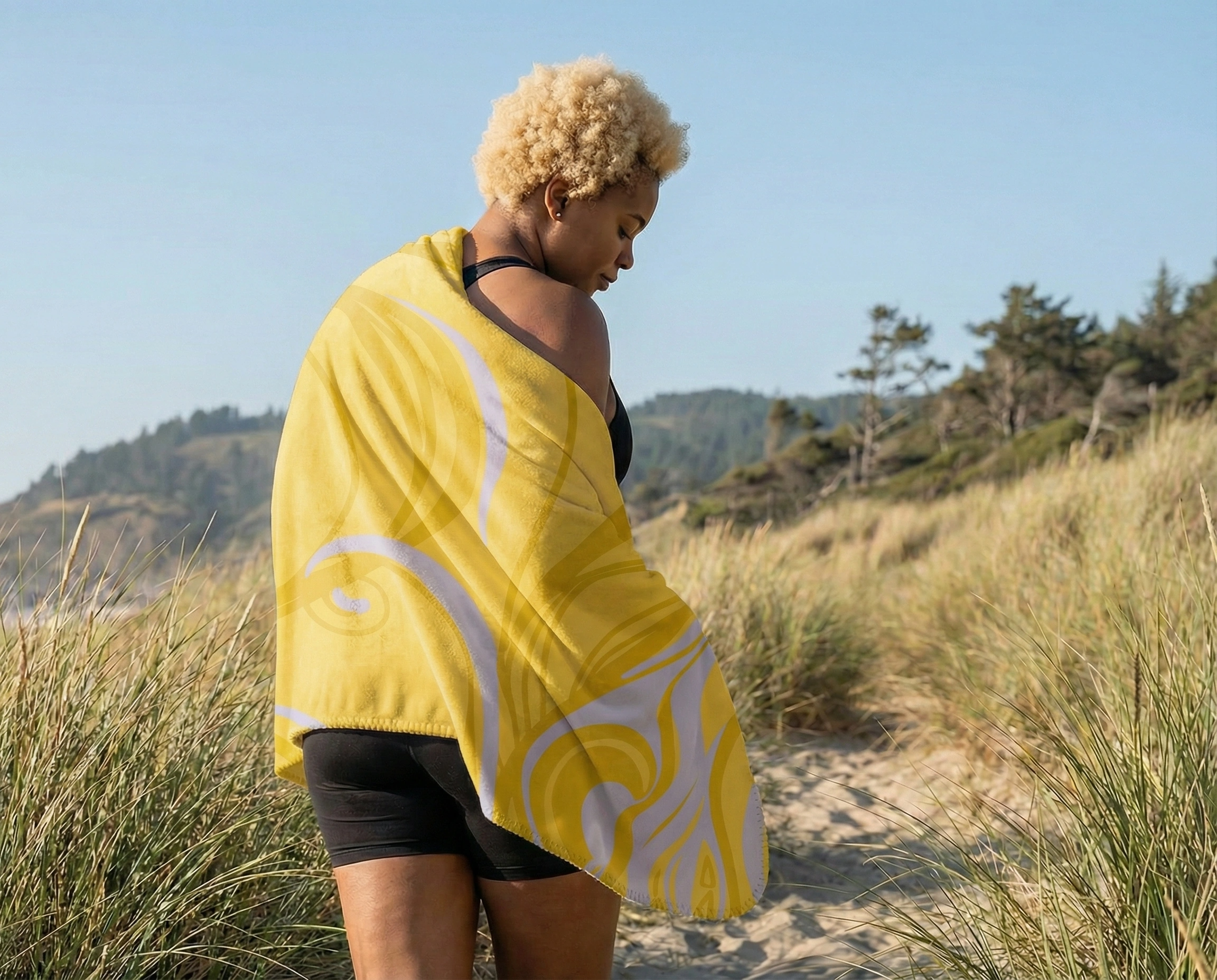 Woman standing on a sandy coastal path, wearing black activewear with a yellow yoga mat featuring subtle swirl patterns draped over her shoulders in a natural outdoor setting.