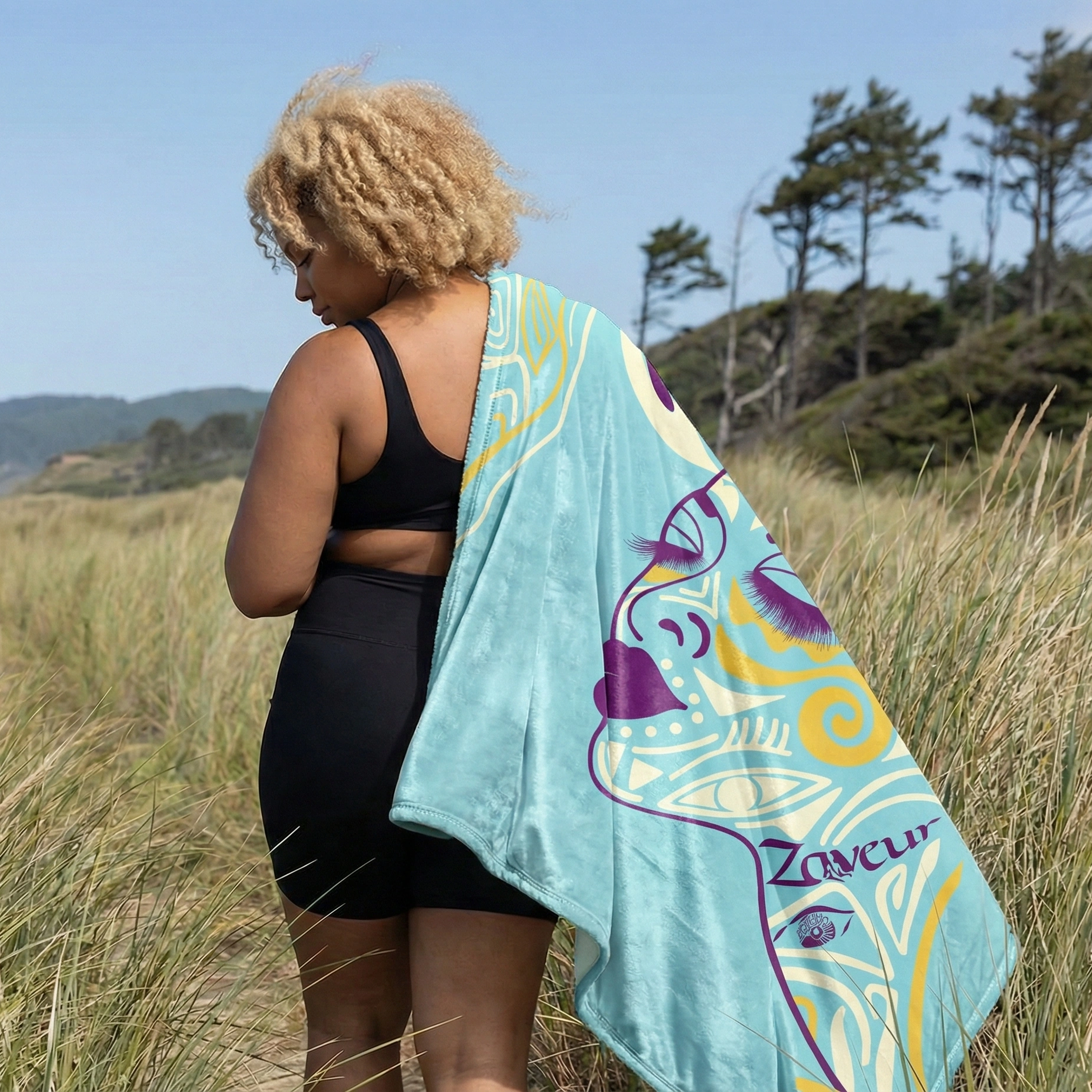 Woman standing outdoors in a grassy coastal landscape wearing black activewear and draped in a cyan abstract throw blanket featuring a stylized face illustration and modern line art design.