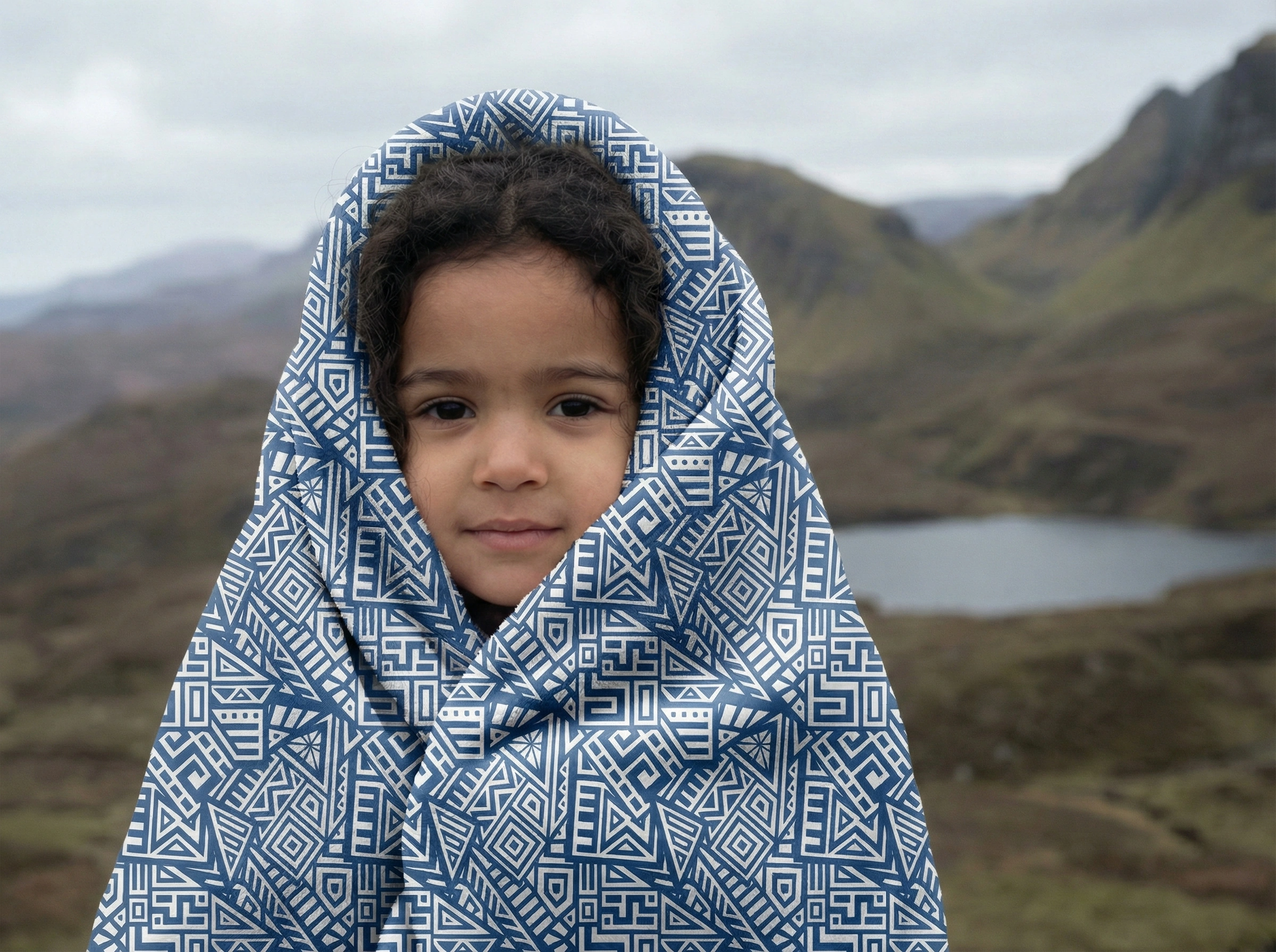 Child wrapped in a blue geometric patterned shawl standing in a mountain landscape, representing warmth, comfort, and mindful lifestyle.