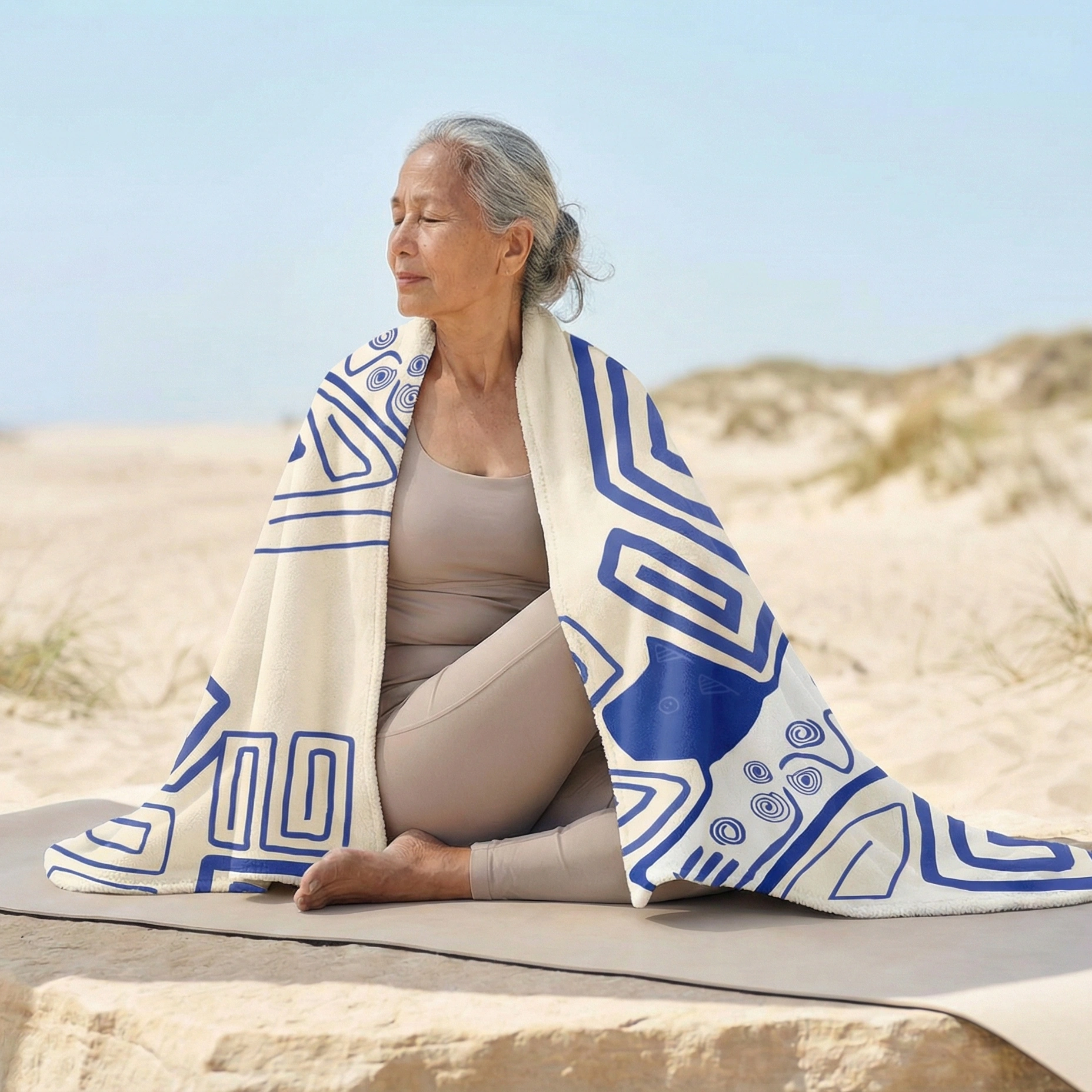Person seated on a yoga mat at the beach, wrapped in a white blanket with blue geometric patterns during a calm meditation pose.